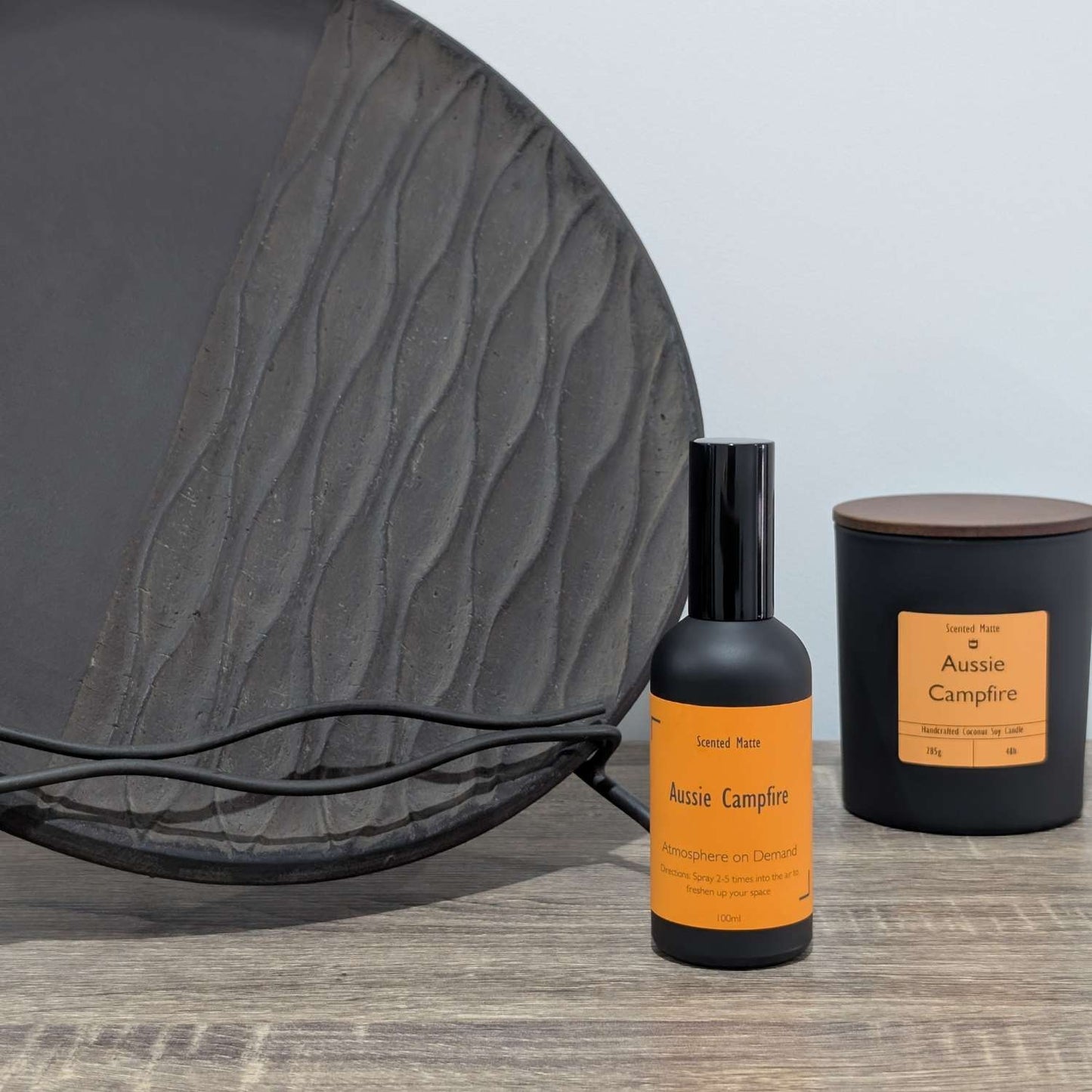 A matte black room spray bottle and a minimalist matte black candle jar, styled next to a large textured charcoal grey decorative plate.