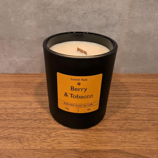 A minimalist matte black candle jar with a wooden wick, sitting on a light oak wood surface against a textured grey background.