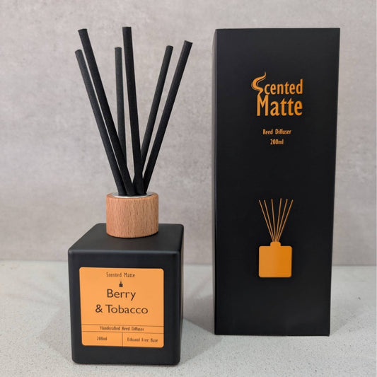 A minimalist matte black reed diffuser with a light wood cap and black reeds, shown next to its black and orange branded box on a grey background.