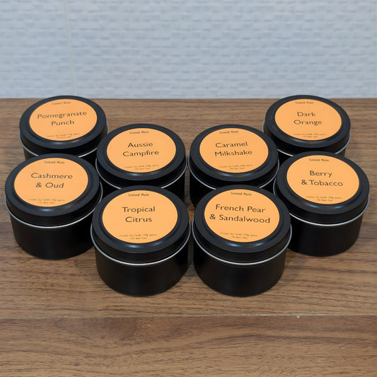Assorted coconut soy candles in black tins with orange labels on a wooden surface.