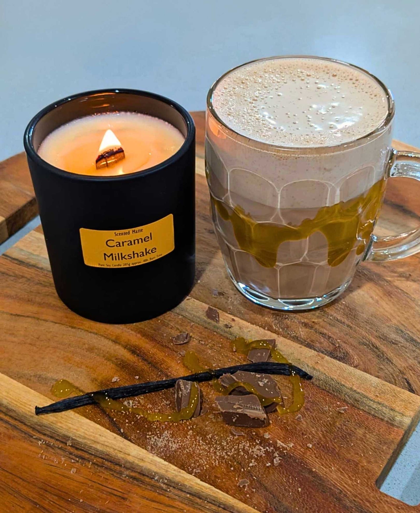 A wooden board with chocolate shavings, a vanilla bean and a milkshake with a lit wooden wick candle in a matte black jar