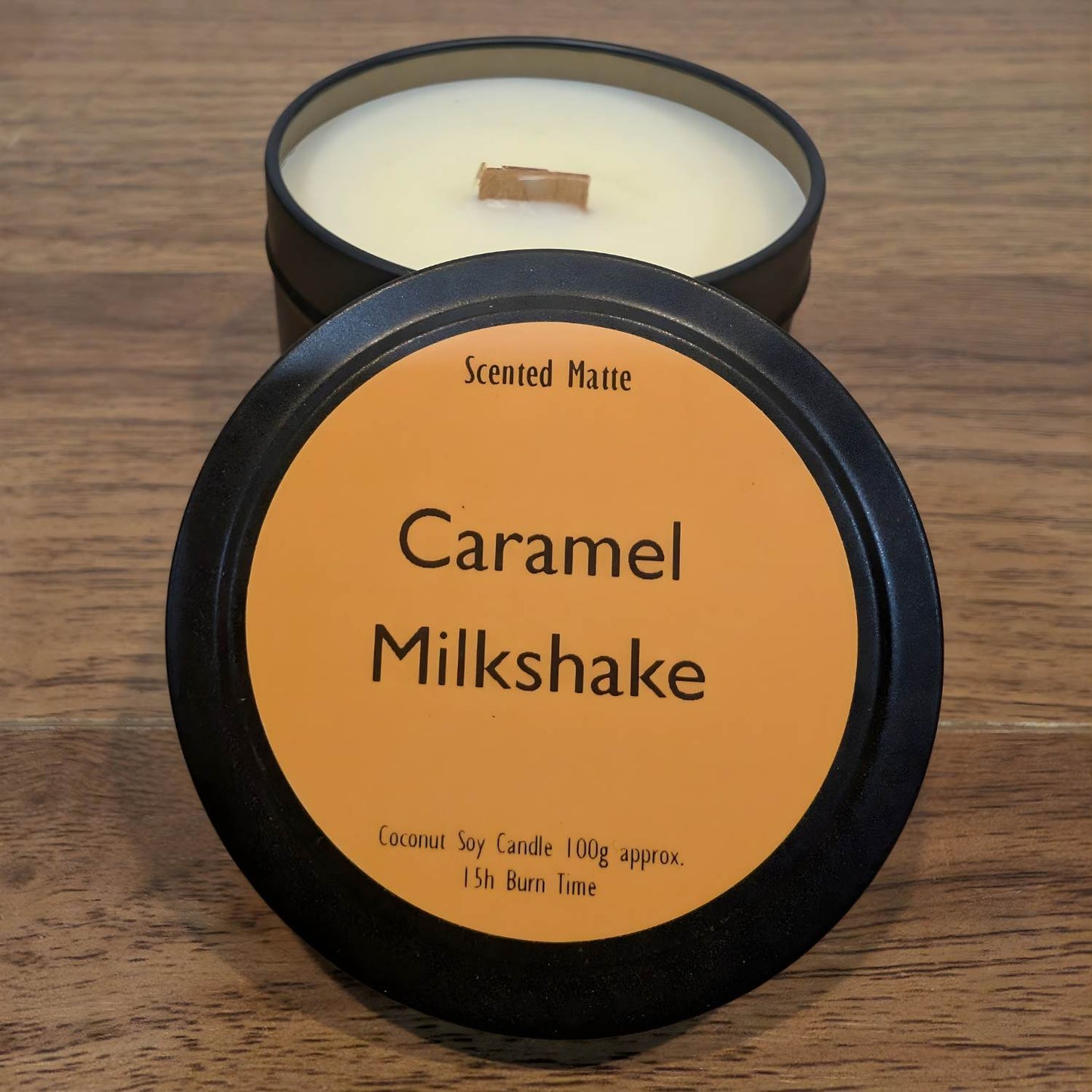 A wooden wick candle in a black tin with orange label on the lid on a wooden surface.