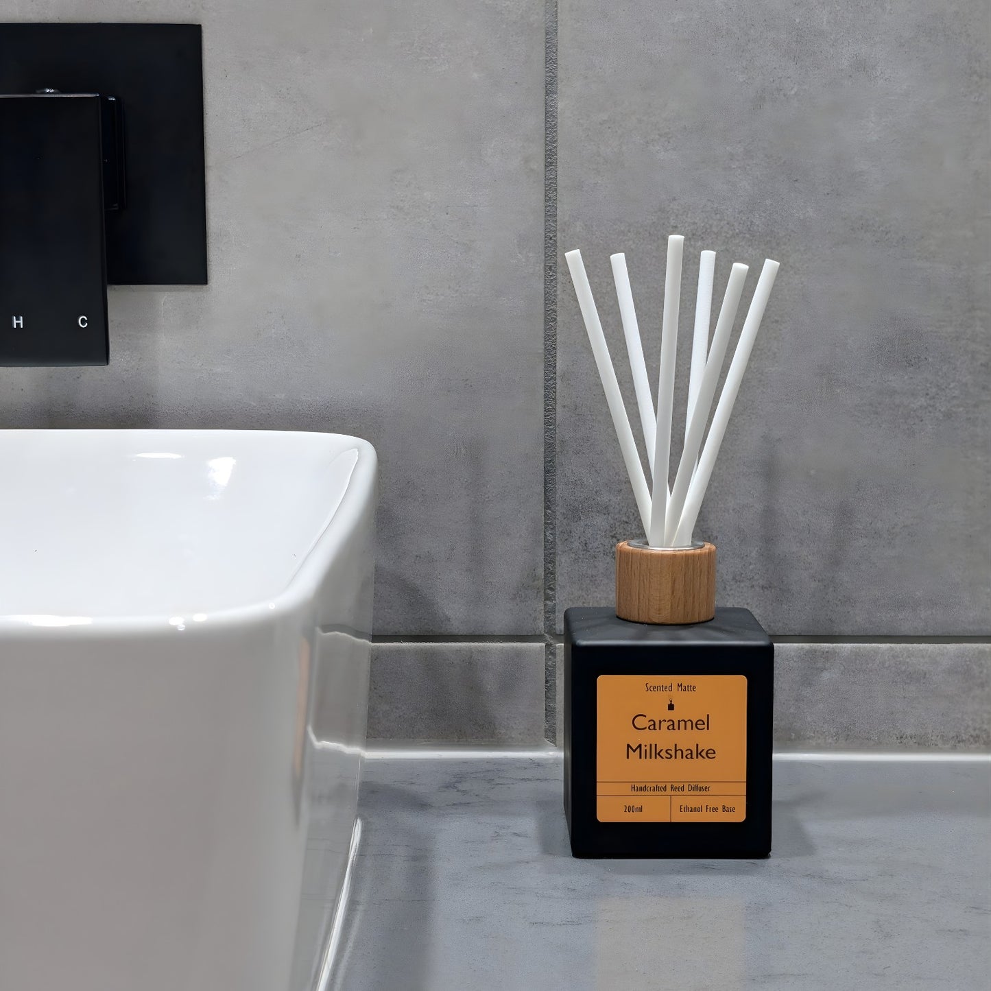 A minimalist matte black reed diffuser with orange label and white fiber reeds, sitting on a grey stone vanity next to a white ceramic sink and grey tiled wall.