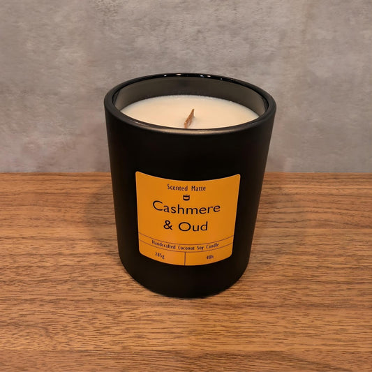 A minimalist matte black candle jar with a wooden wick, sitting on a light oak wood surface against a textured grey background.