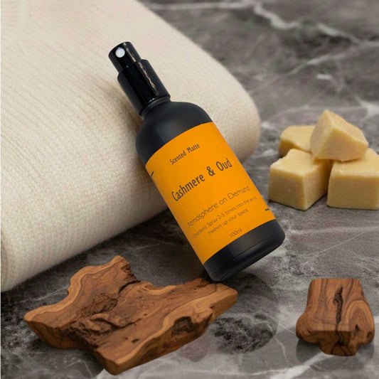 A minimalist matte black room spray bottle with a vibrant orange label, nestled in soft cream knitted fabric with dark oud wood pieces.
