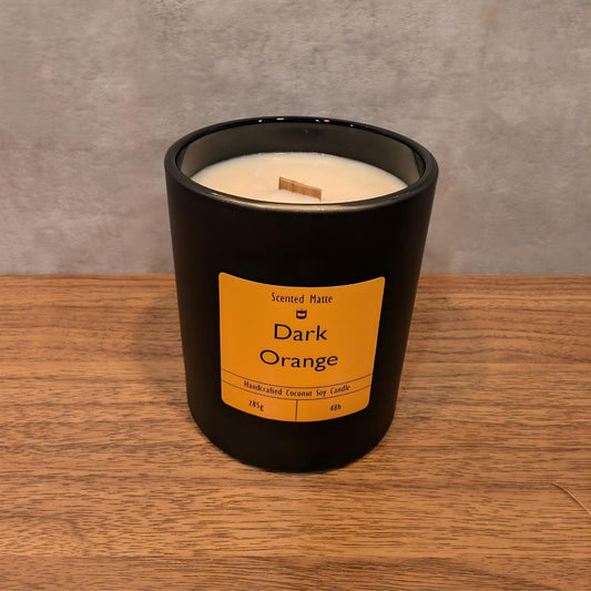 A minimalist matte black candle jar with a wooden wick, sitting on a light oak wood surface against a textured grey background.