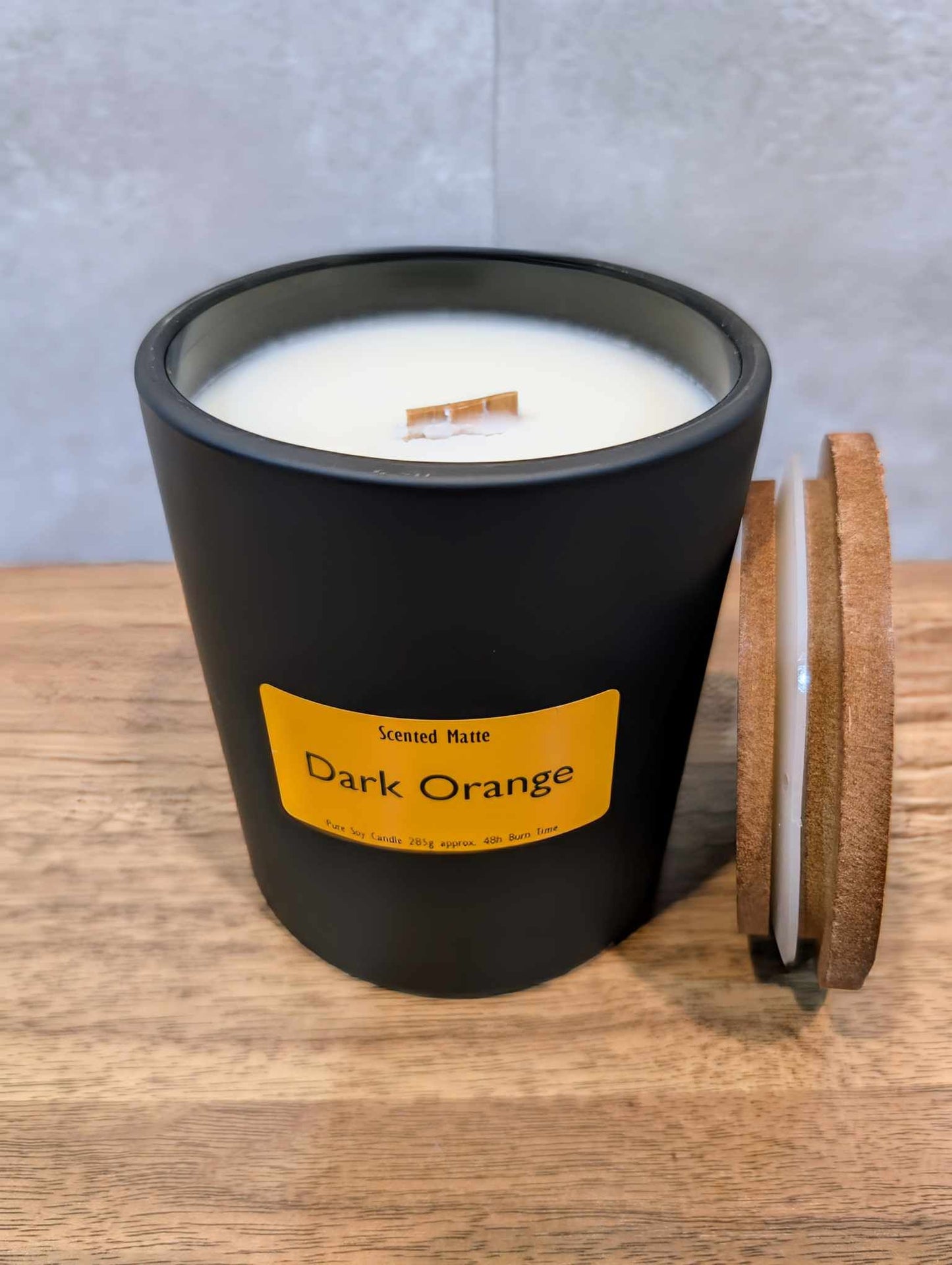 A soy wooden wick candle in a matte black glass jar with an orange label and wooden lid