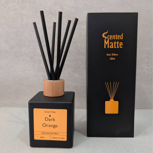 A minimalist matte black reed diffuser with a light wood cap and black reeds, shown next to its black and orange branded box on a grey background.