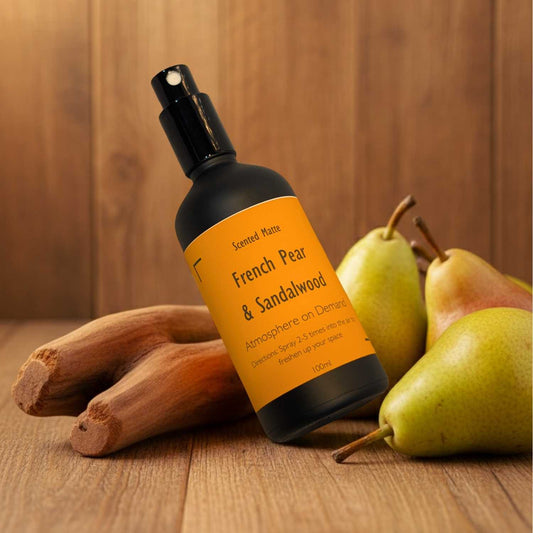 A minimalist matte black room spray bottle with a vibrant orange label, sitting next to a green pear and light sandalwood.