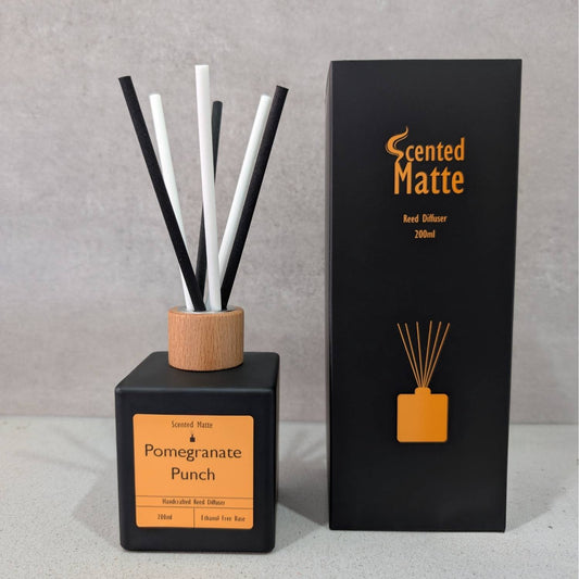 A minimalist matte black reed diffuser with a light wood cap and black/white reeds, shown next to its black and orange branded box on a grey background.