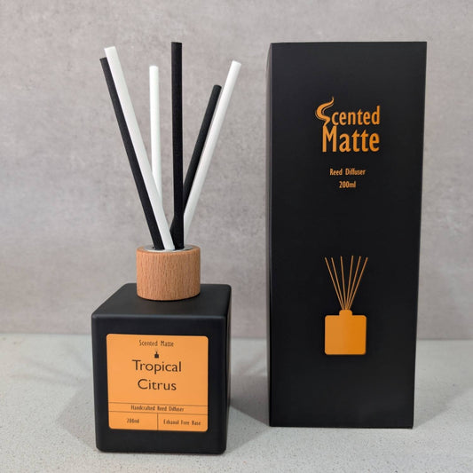 A minimalist matte black reed diffuser with a light wood cap and black/white reeds, shown next to its black and orange branded box on a grey background.