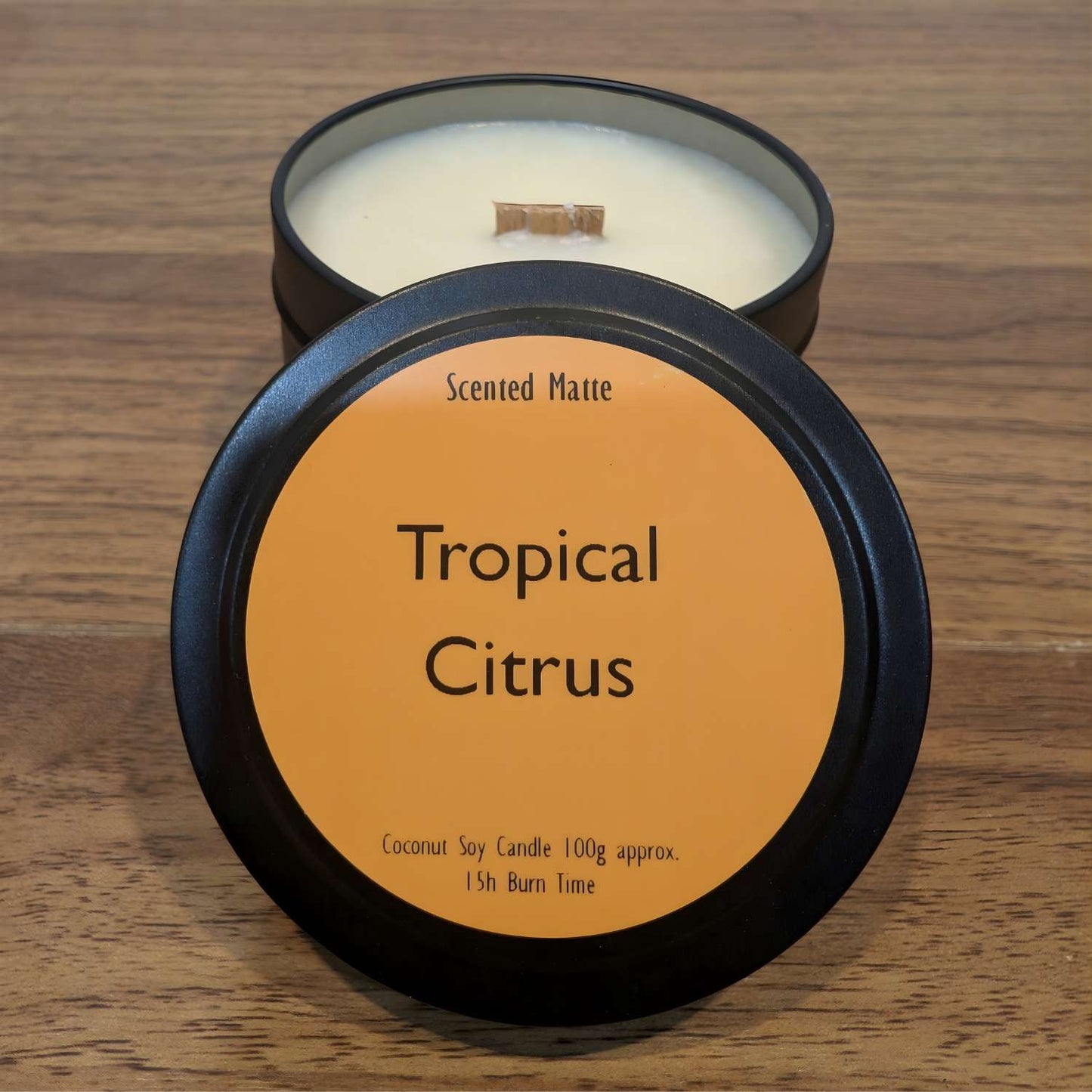 A wooden wick candle in a black tin with orange label on the lid on a wooden surface.
