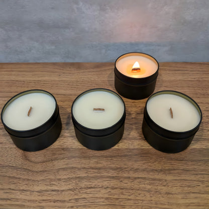 Four wooden wick candles in black tins with white wax laid out in a line on a wooden surface, one of which is lit.