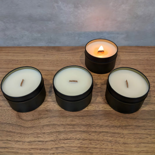Four wooden wick candles in black tins with white wax laid out in a line on a wooden surface, one of which is lit.