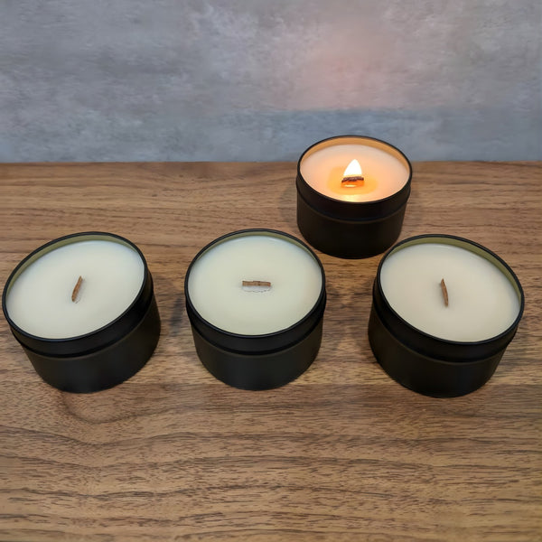 Four wooden wick candles in black tins with white wax laid out in a line on a wooden surface, one of which is lit.