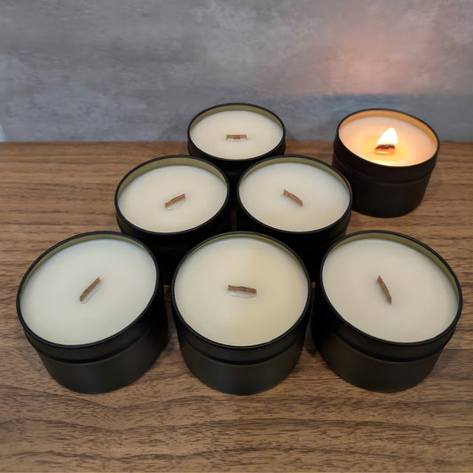 7 wooden wick candles in black tins with white wax on a wooden surface laid out in a pyramid, one of which is lit.