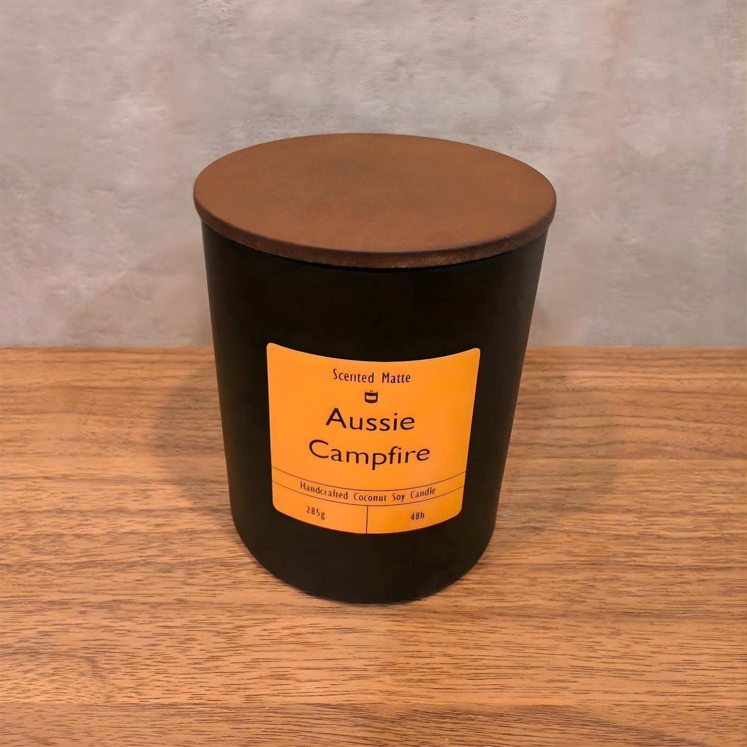 A minimalist matte black candle jar with a vibrant orange label and a dark wood lid.