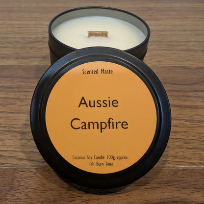 A wooden wick candle in a black tin with orange label on the lid on a wooden surface.
