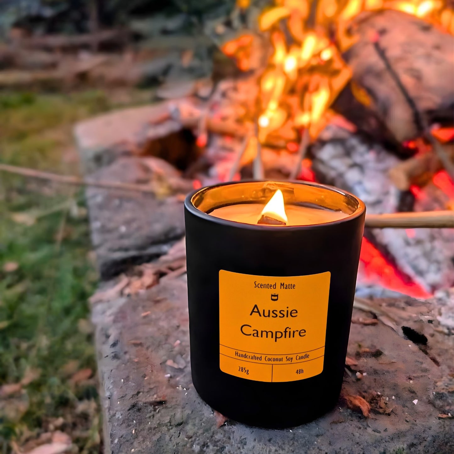 A minimalist matte black candle jar with a wooden wick on stone in front of a crackling wood fire with dried leaves.