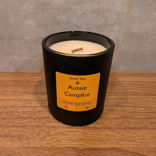 A minimalist matte black candle jar with a wooden wick, sitting on a light oak wood surface against a textured grey background.