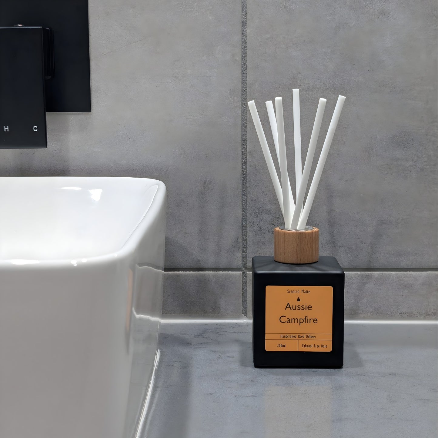 A minimalist matte black reed diffuser with orange label, wooden cap and white fiber reeds, sitting on a grey stone vanity next to a white ceramic sink and grey tiled wall.