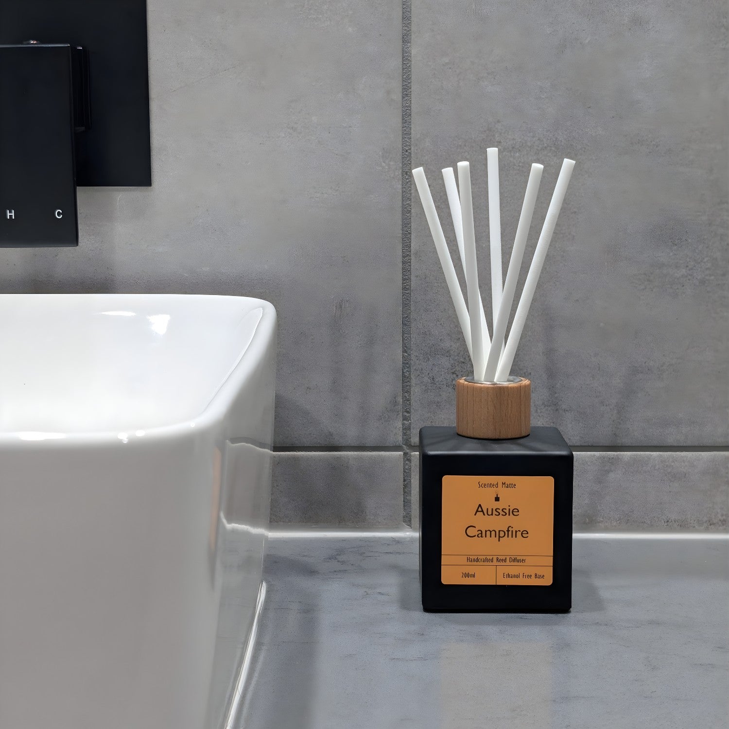 A minimalist matte black reed diffuser with orange label, wooden cap and white fiber reeds, sitting on a grey stone vanity next to a white ceramic sink and grey tiled wall.