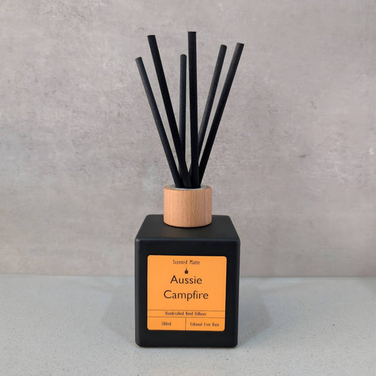 A minimalist matte black square reed diffuser with an orange label, light wood cap and six black fiber reeds, set against a grey background.
