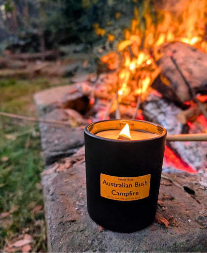 A lit wooden wick candle on a stone with a campfire and trees in the background