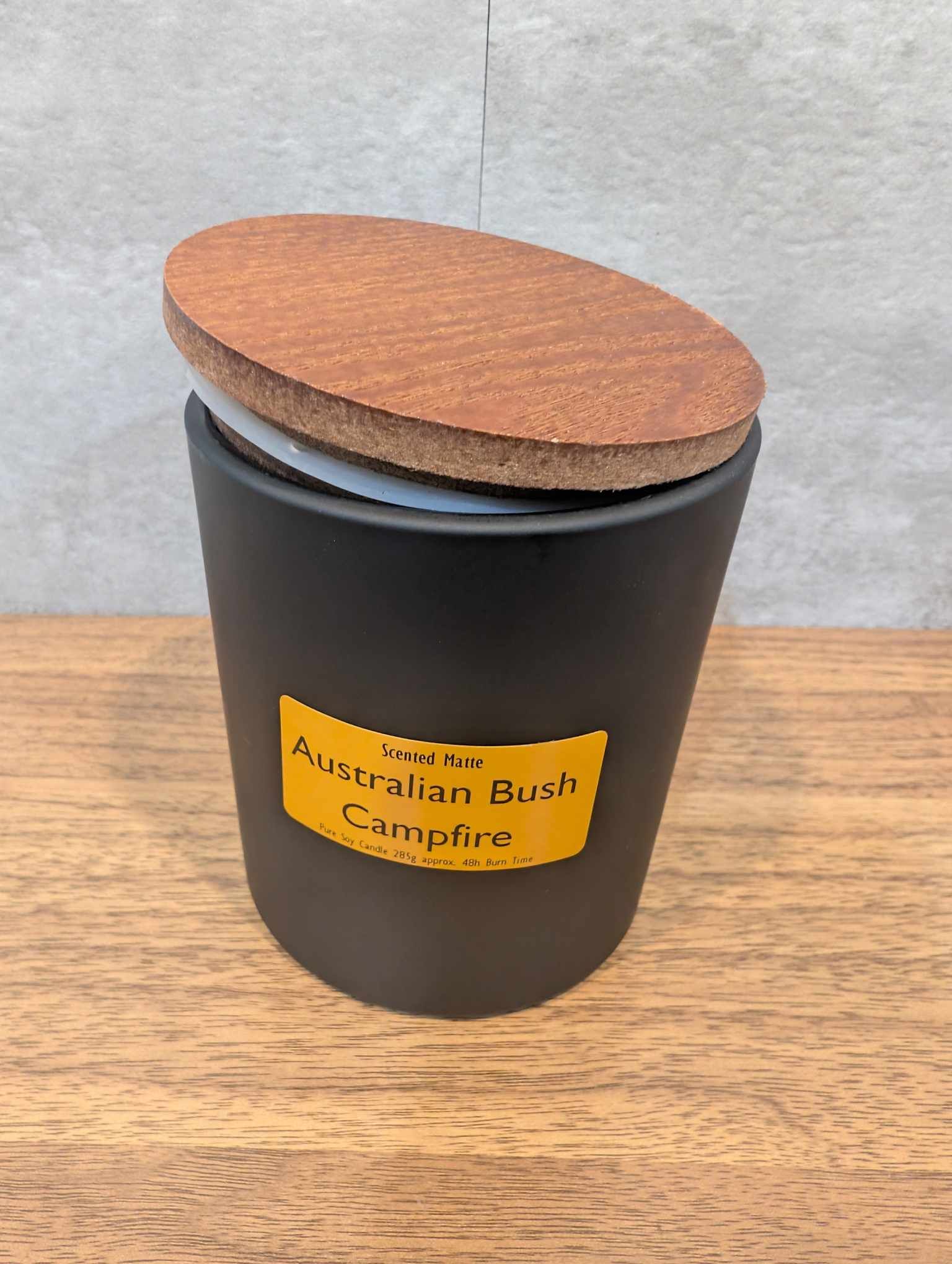 A matte black candle jar with orange label and a wooden lid on top