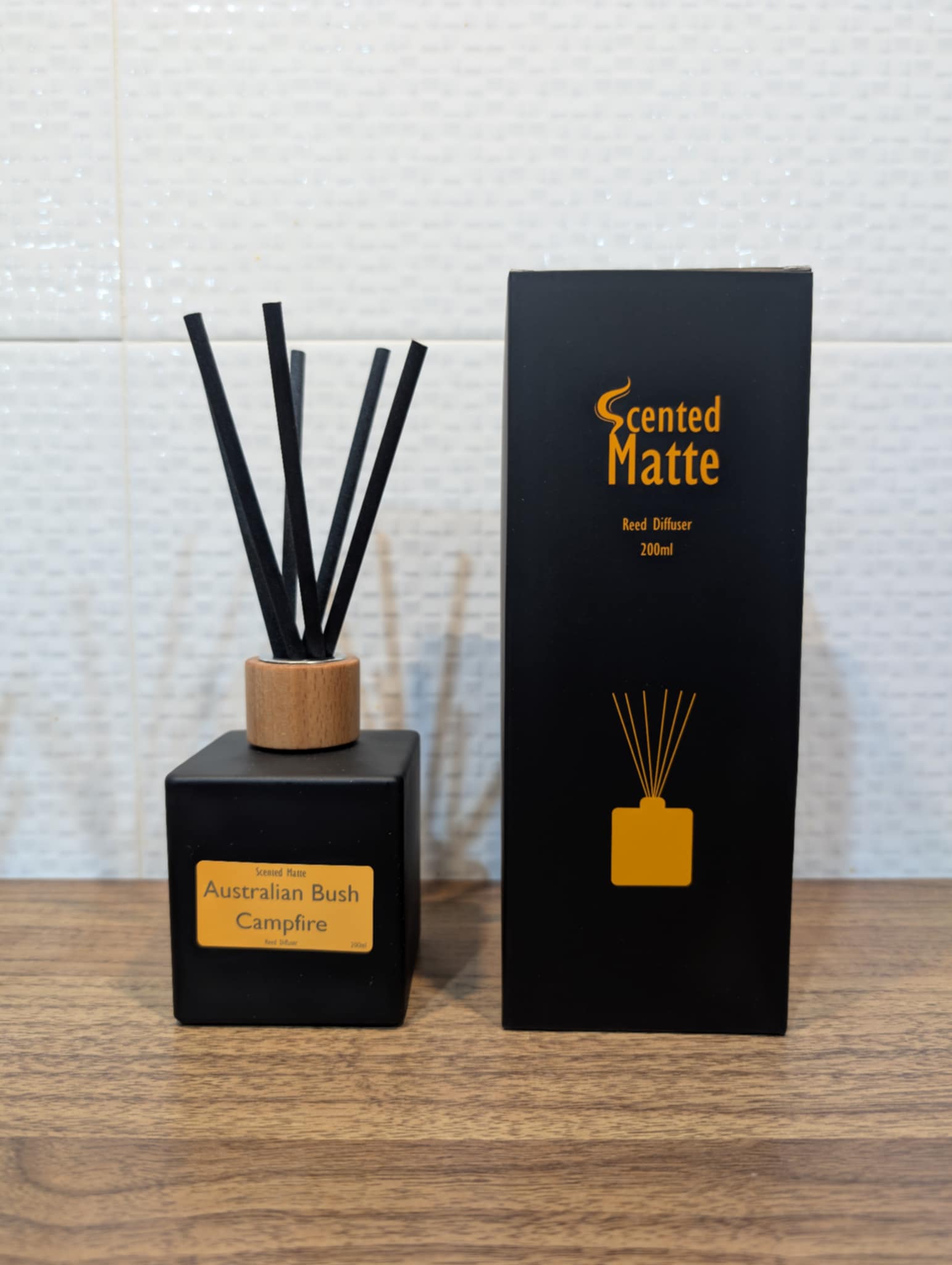 Matte black diffuser bottle with black reed sticks and wooden cap, next to its packaging on a wooden surface.