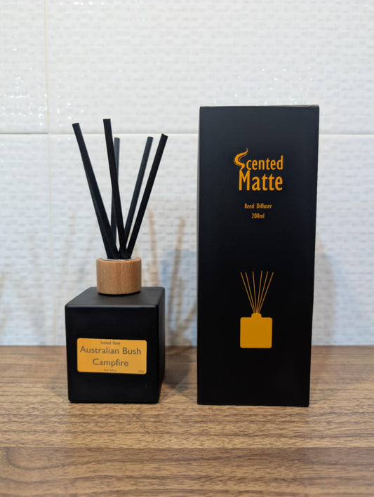 Matte black diffuser bottle with black reed sticks and wooden cap, next to its packaging on a wooden surface.