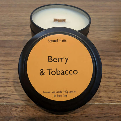 A wooden wick candle in a black tin with orange label on the lid on a wooden surface.