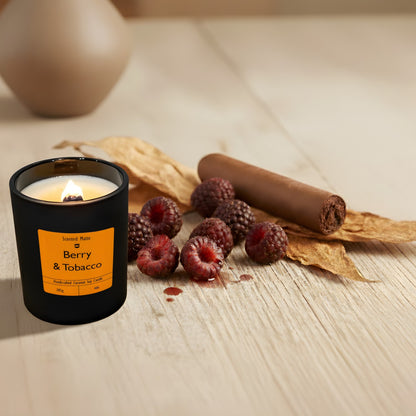 A minimalist matte black candle jar with a lit wooden wick, beside dark red berries and dried tobacco leaves on a light wooden surface.
