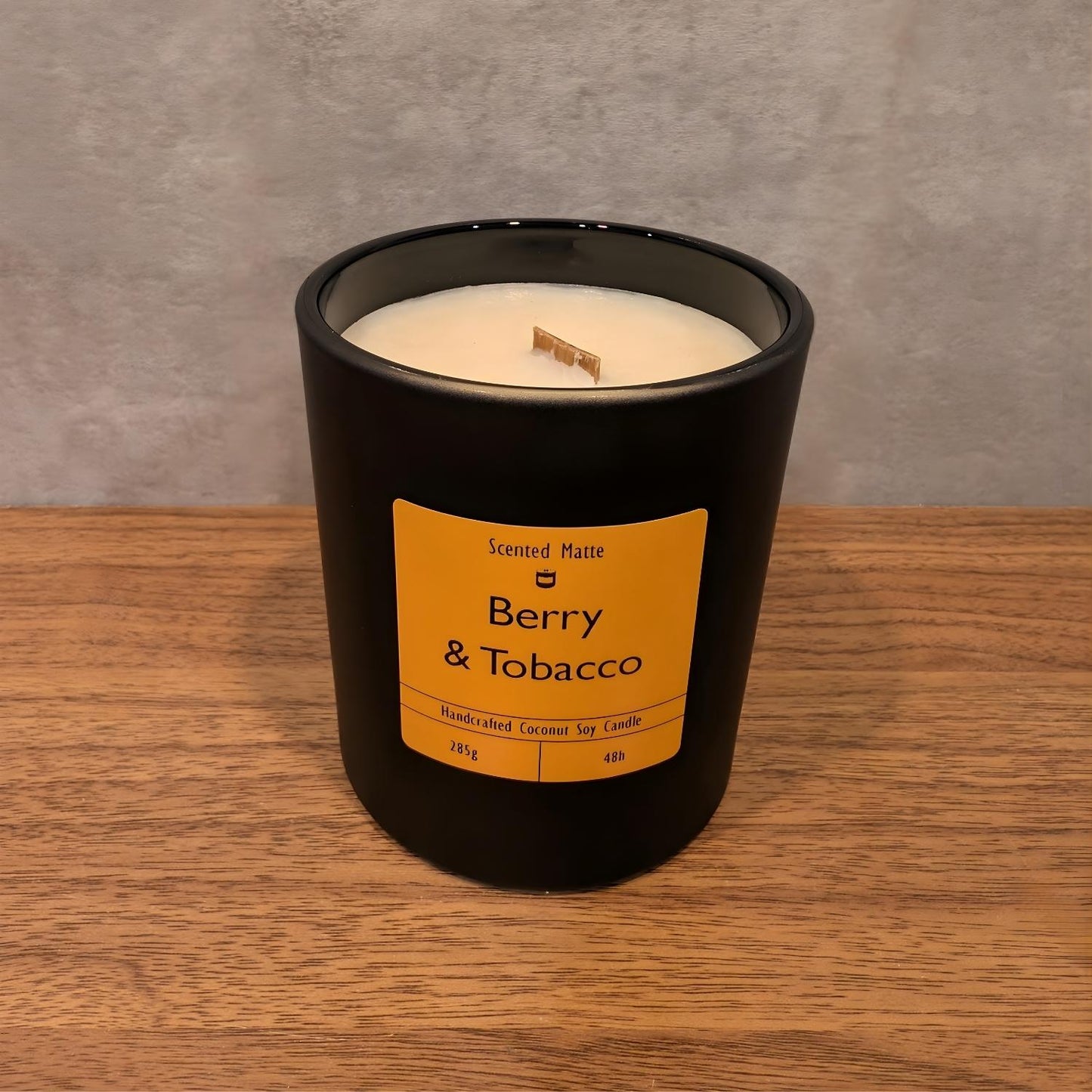 A minimalist matte black candle jar with a wooden wick, sitting on a light oak wood surface against a textured grey background.