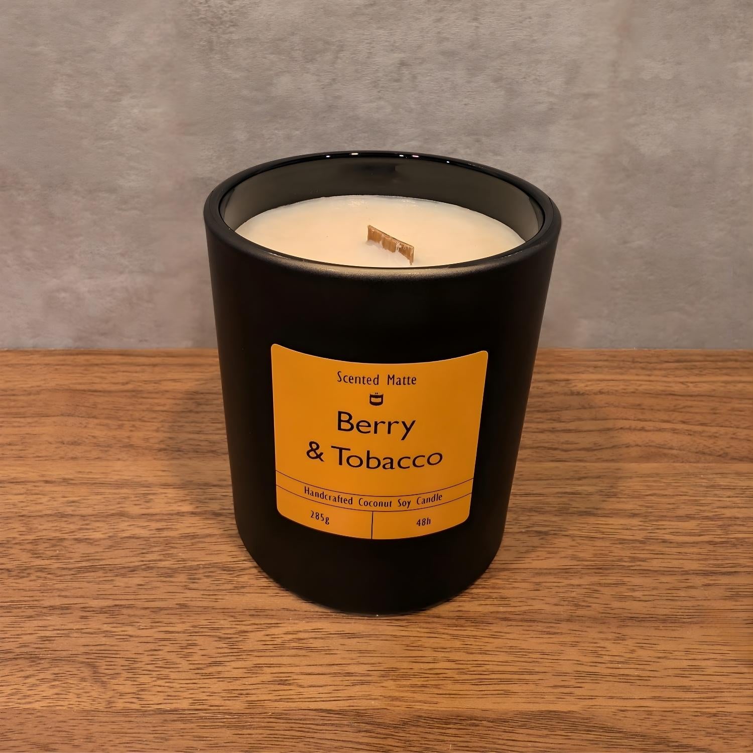 A minimalist matte black candle jar with a wooden wick, sitting on a light oak wood surface against a textured grey background.