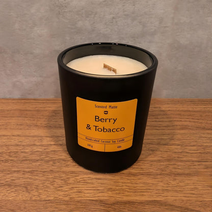 A minimalist matte black candle jar with a wooden wick, sitting on a light oak wood surface against a textured grey background.