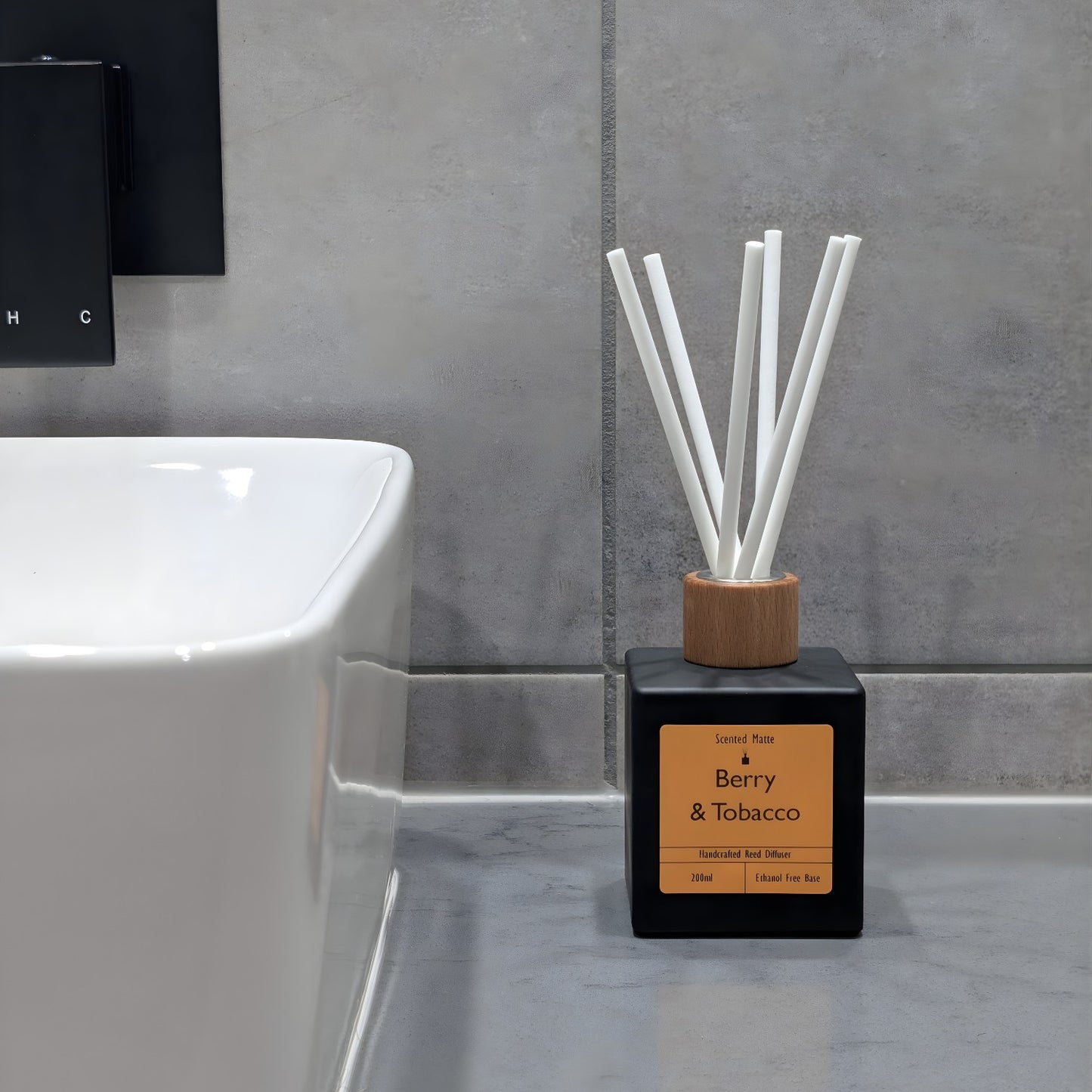 A minimalist matte black reed diffuser with orange label and white fiber reeds, sitting on a grey stone vanity next to a white ceramic sink and grey tiled wall.