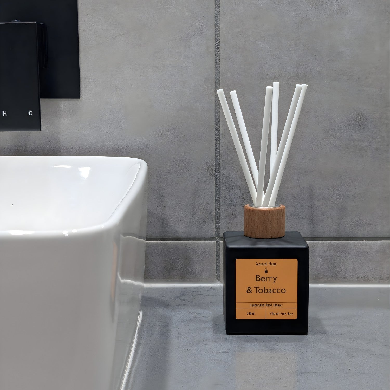 A minimalist matte black reed diffuser with orange label and white fiber reeds, sitting on a grey stone vanity next to a white ceramic sink and grey tiled wall.