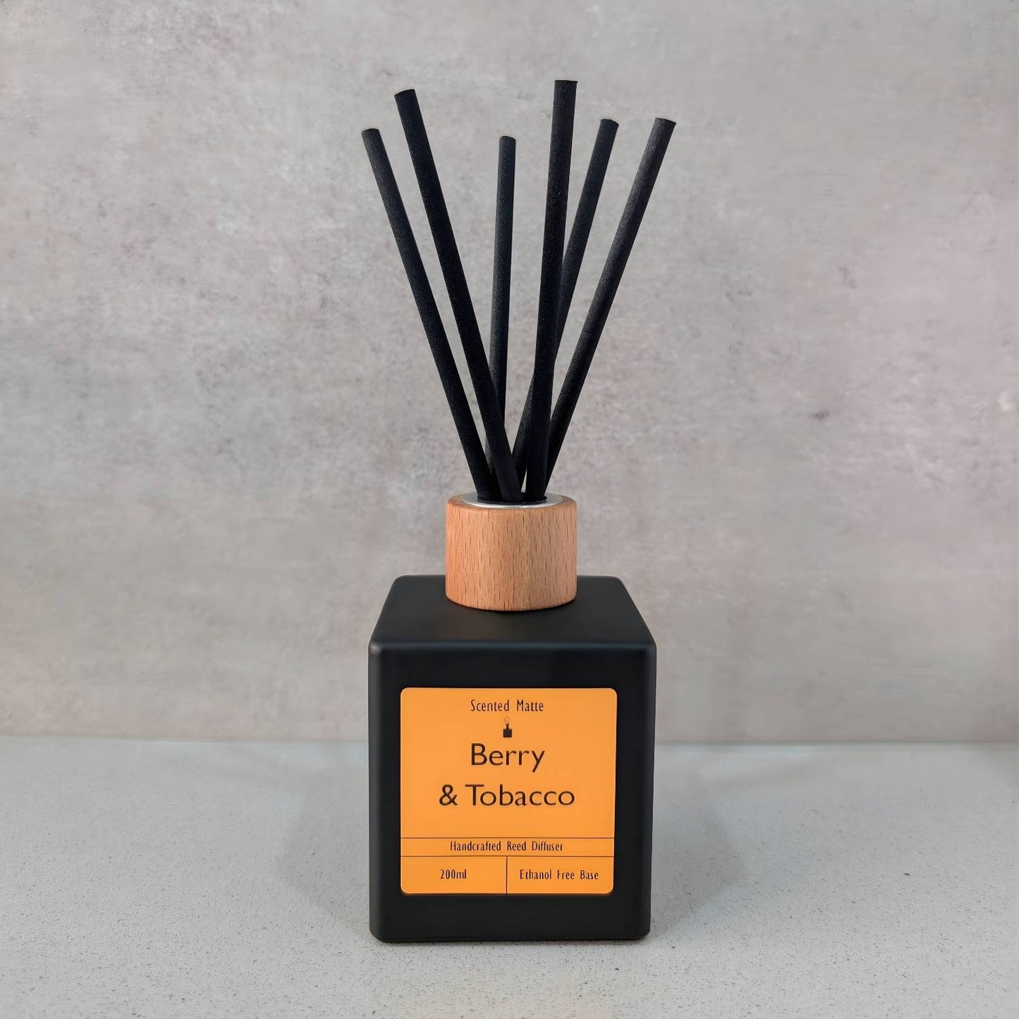 A minimalist matte black square reed diffuser with an orange label, light wood cap and six black fiber reeds, set against a grey background.