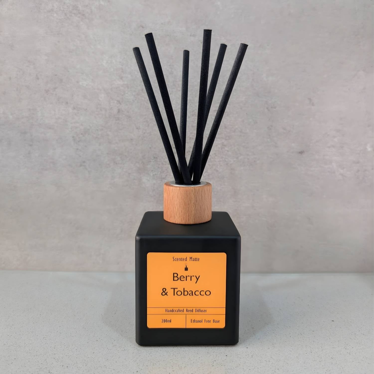 A minimalist matte black square reed diffuser with an orange label, light wood cap and six black fiber reeds, set against a grey background.
