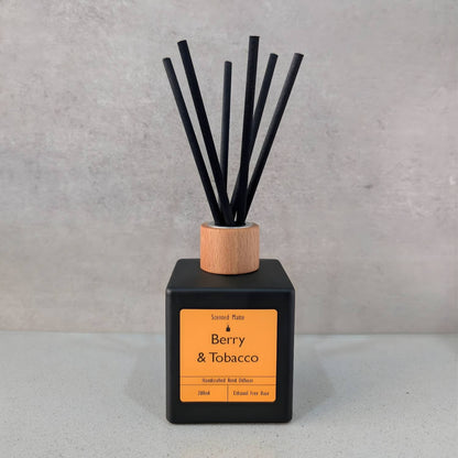 A minimalist matte black square reed diffuser with an orange label, light wood cap and six black fiber reeds, set against a grey background.