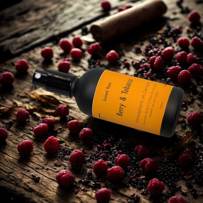 A minimalist matte black room spray bottle with a vibrant orange label, styled next to dark red berries and dried tobacco leaves on a shadowed surface.