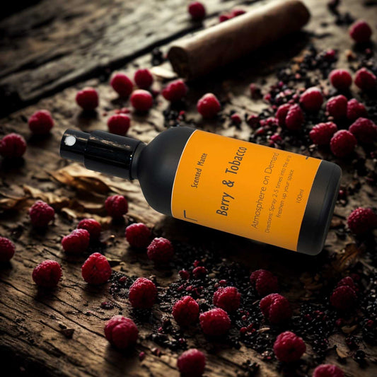 A minimalist matte black room spray bottle with a vibrant orange label, styled next to dark red berries and dried tobacco leaves on a shadowed surface.