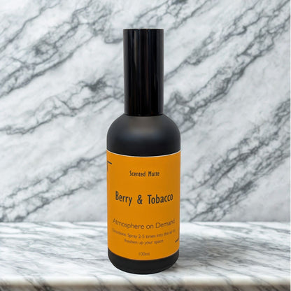 A minimalist matte black 100ml room spray bottle with a vibrant orange label, centered on a white marble surface.