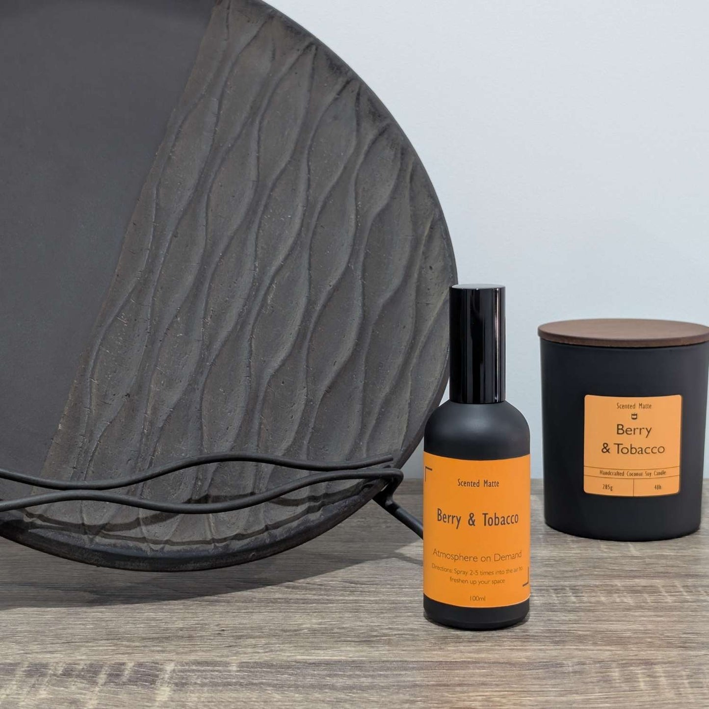 A matte black room spray bottle and a minimalist matte black candle jar, styled next to a large textured charcoal grey decorative plate.