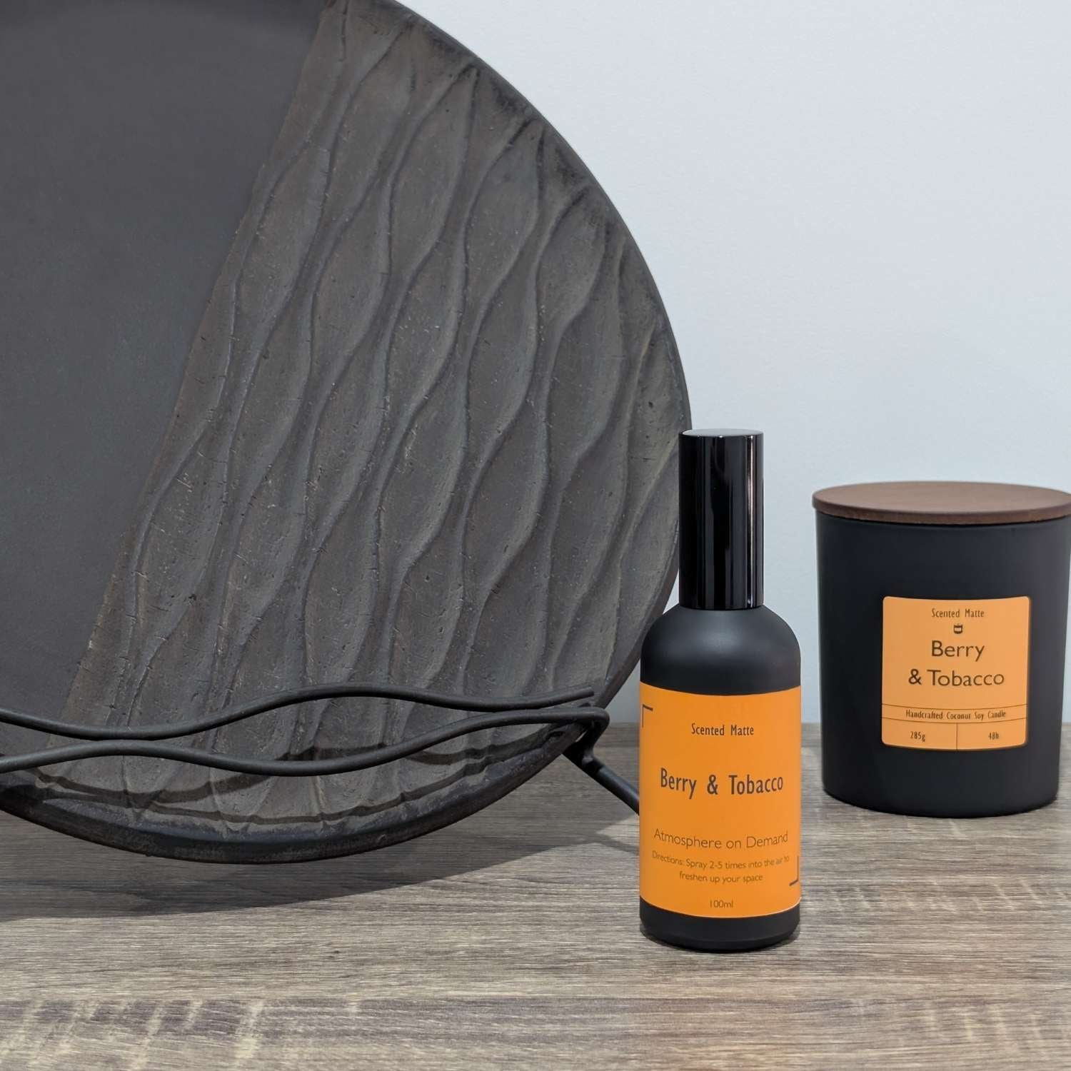A matte black room spray bottle and a minimalist matte black candle jar, styled next to a large textured charcoal grey decorative plate.