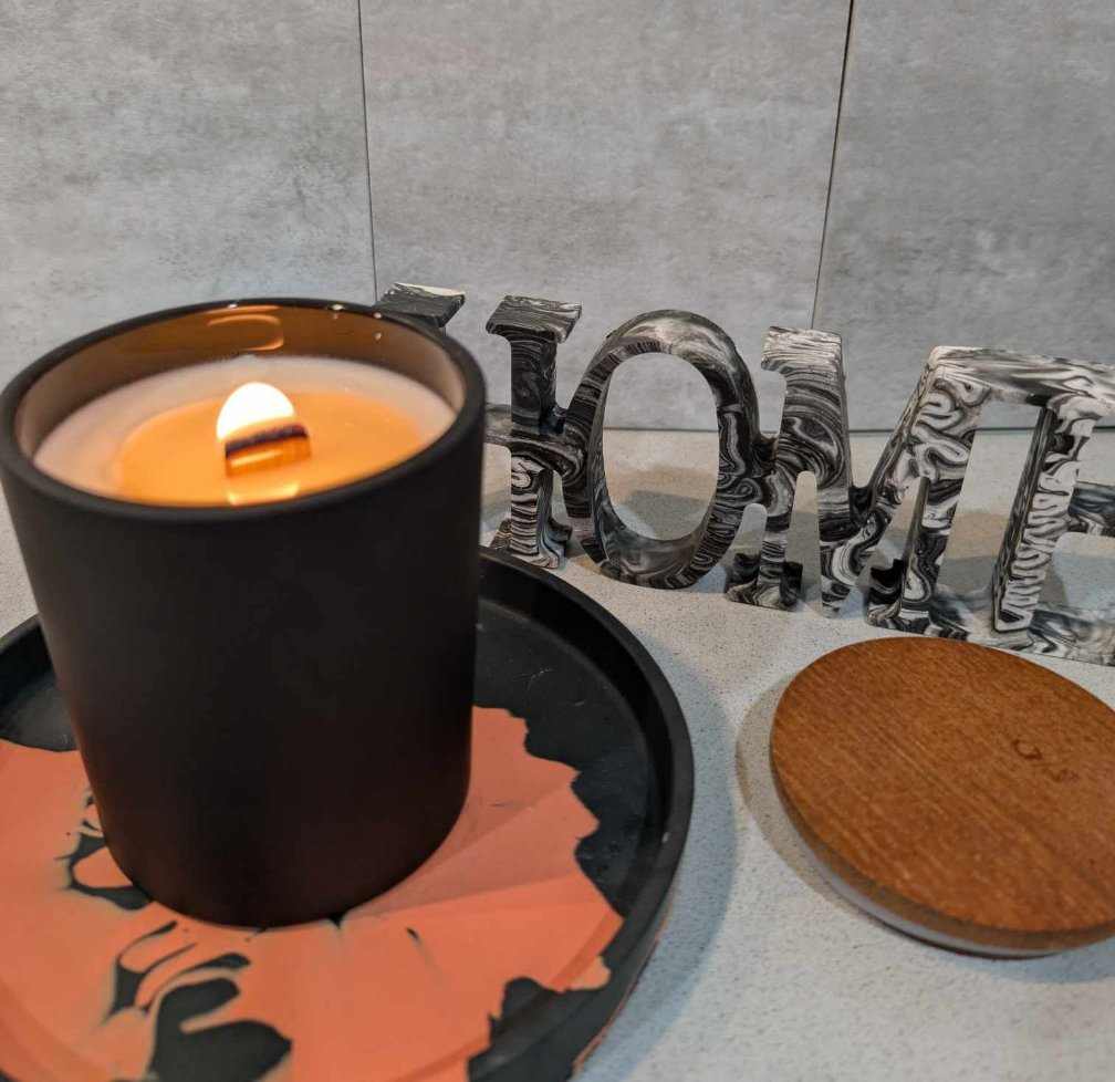 A lit soy wooden wck candle in a matte black glass jar with decorative items