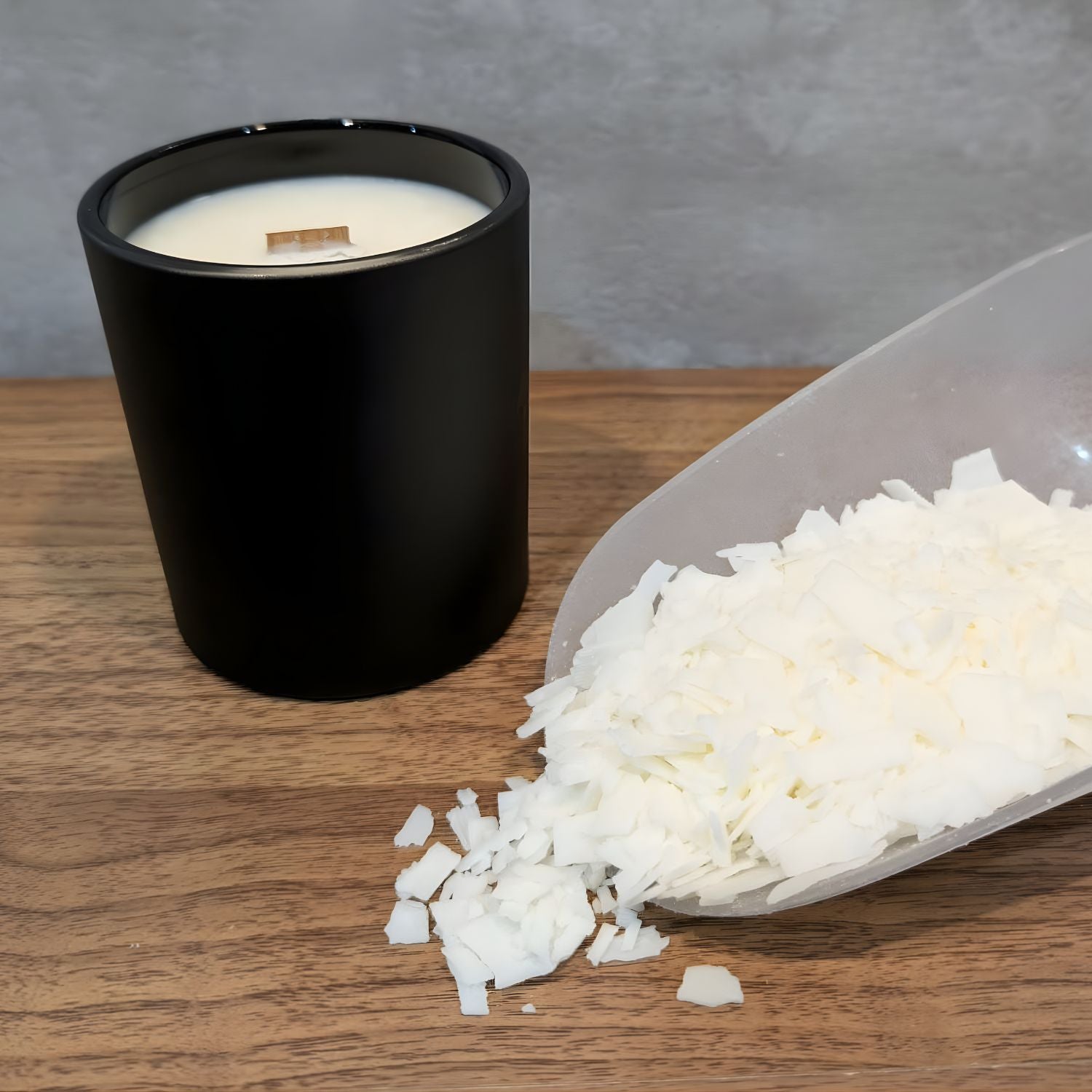 A minimalist matte black candle jar with a wooden wick beside a scoop of flaked wax.