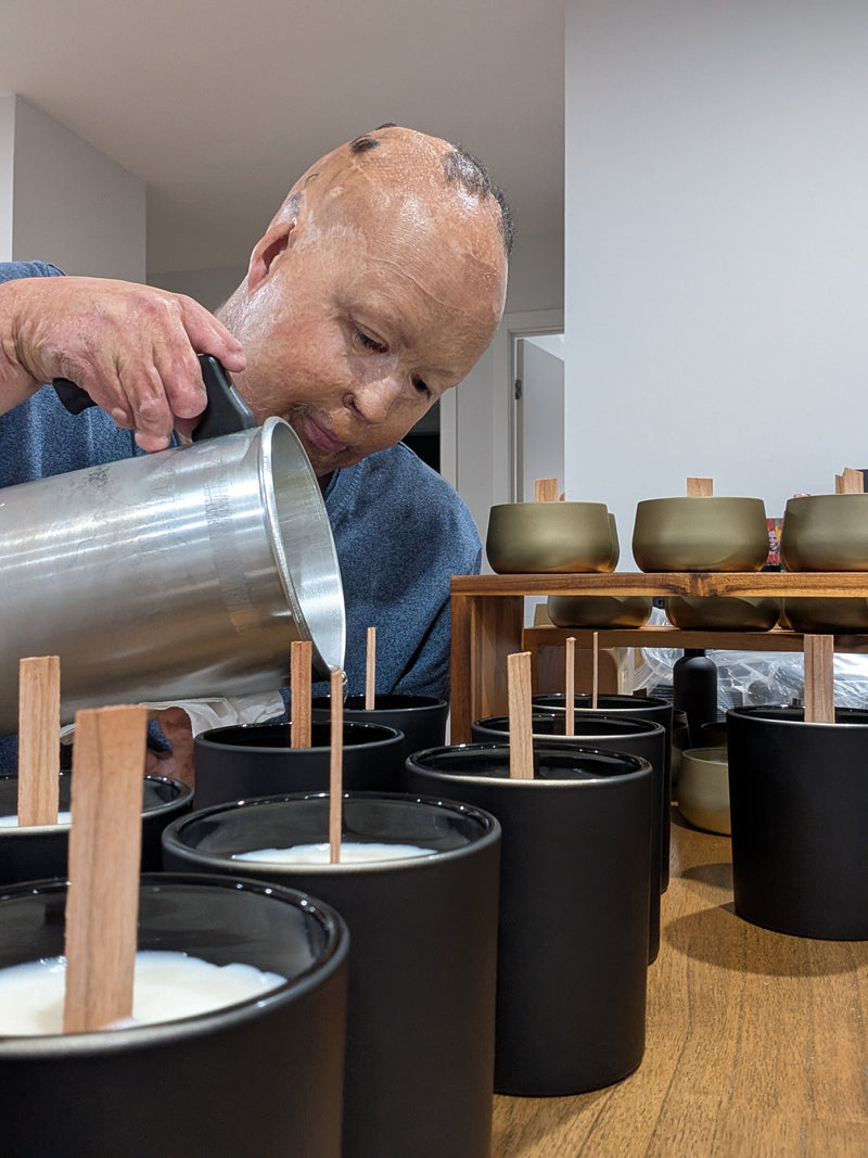 Person pouring liquid into matte black containers with wooden sticks, surrounded by similar containers and a table in the background.