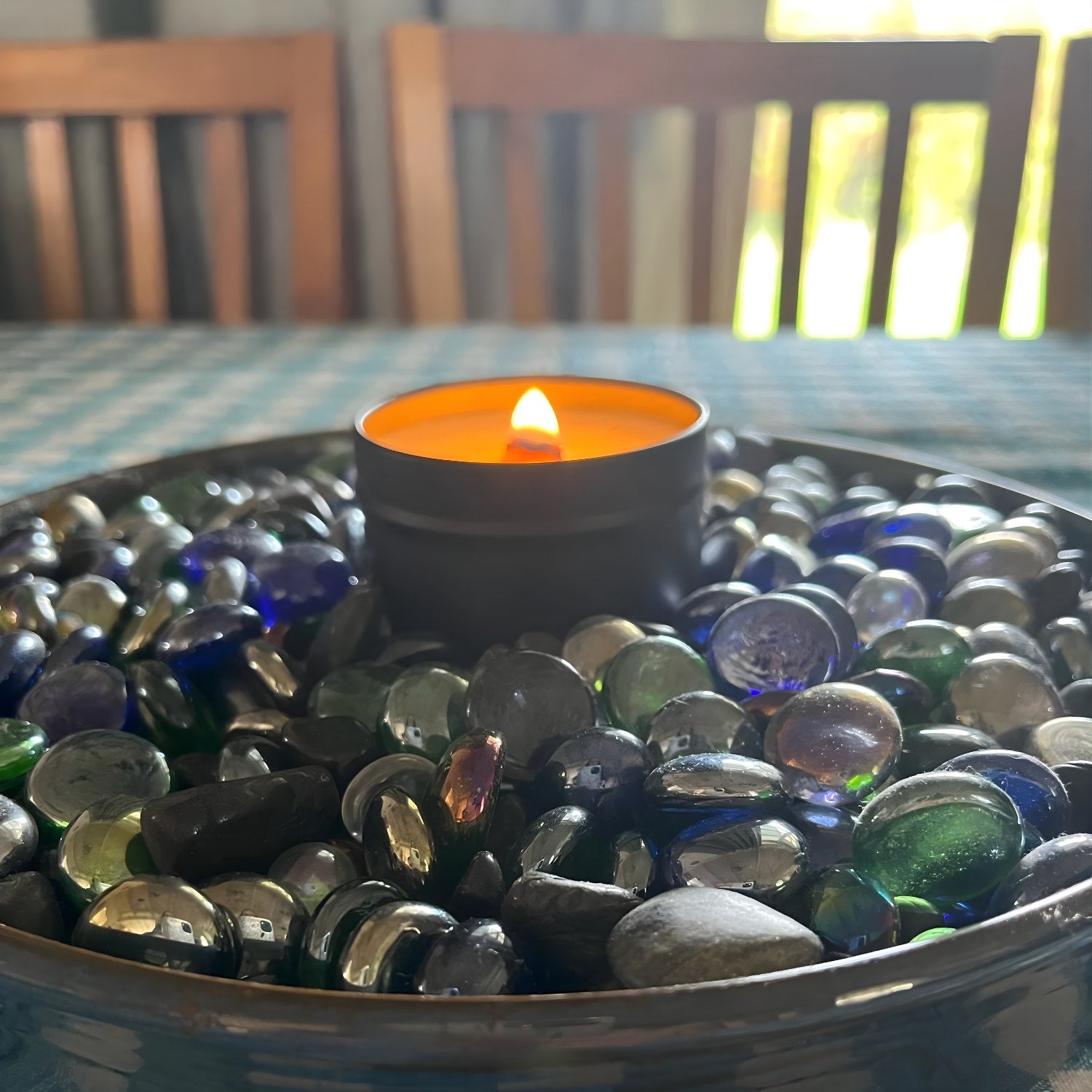Flickering  wooden wick candle in a black tin amounst a bowl of multicolored glass marbles on a table.